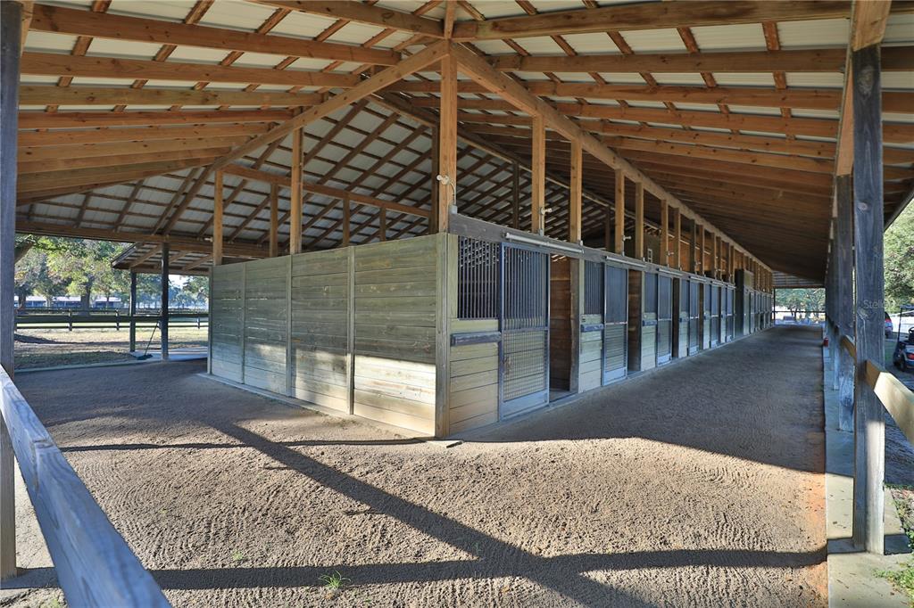 An open-air horse barn features a dirt aisle and multiple wooden stalls with metal doors — 7505 SW 73RD STREET, OCALA, FL