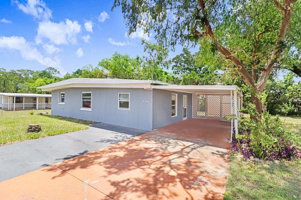 A gray single-story house with a flat roof and an attached carport sits on grass — 39038 HEATH DRIVE, ZEPHYRHILLS, FL
