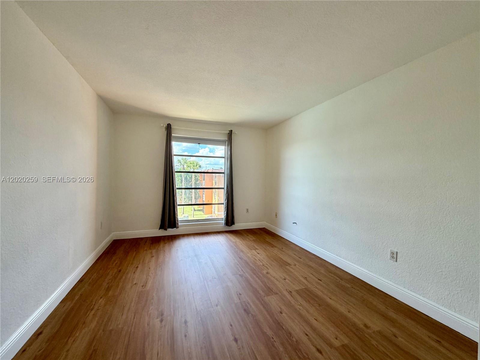 An empty room features wood-look flooring, white walls, and a single window with curtains — 9429 SW 76th St # W38, Miami FL 33173, MIAMI, FL