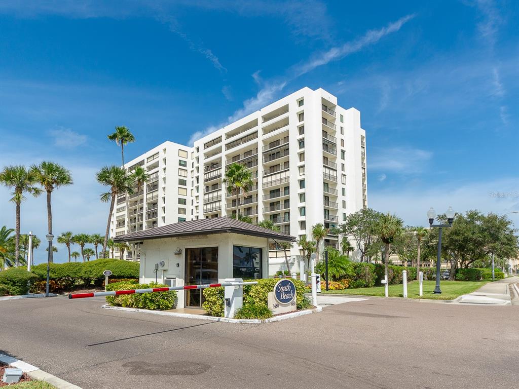 A multi-story white apartment building stands behind a gated security entrance with palm trees — 1460 GULF BOULEVARD UNIT 302, CLEARWATER, FL