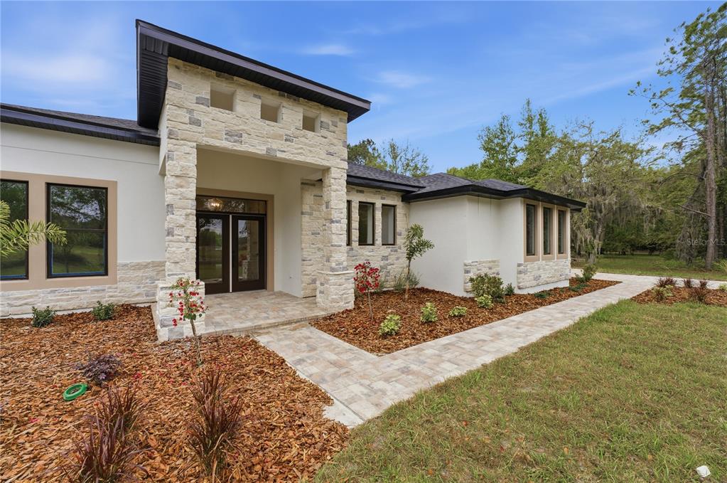 A modern house exterior features a stone-accented entryway, dark-framed windows, and a paved walkway — 1516 PINE ISLAND ROAD, KISSIMMEE, FL