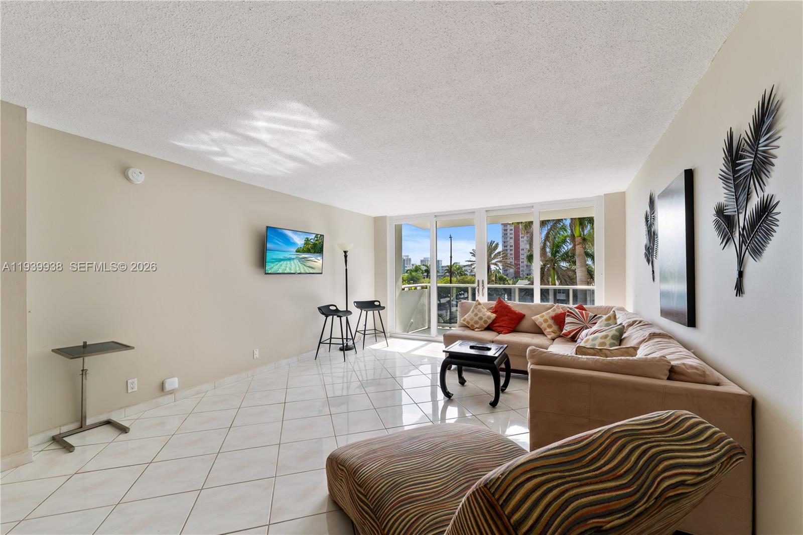 A living room features a beige sectional, tiled floors, a wall-mounted television, and balcony access — 3140 S Ocean Dr # 212, Hallandale Beach FL 33009, HALLANDALE BEACH, FL