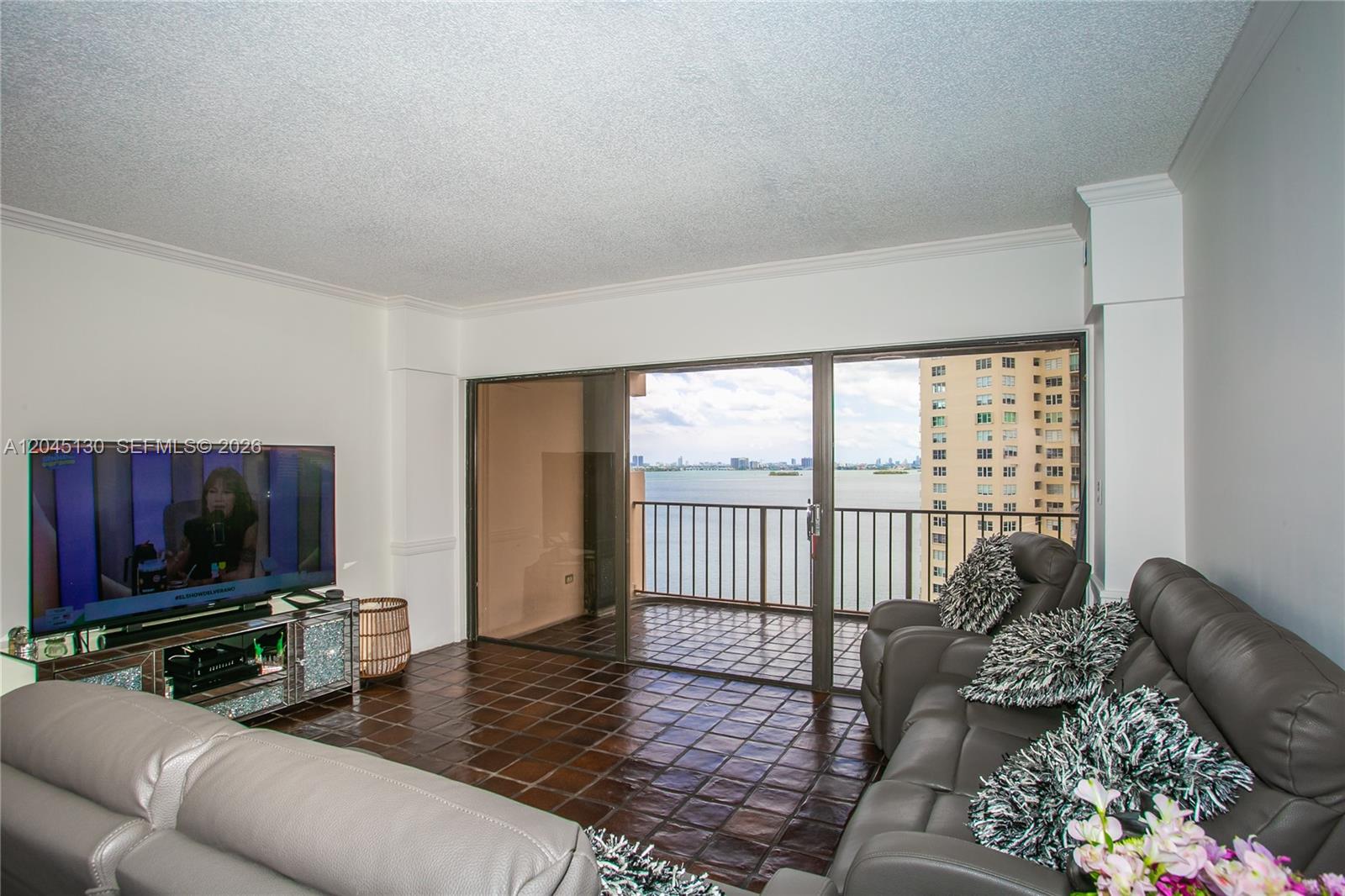 A living room features dark tile floors, a television, and a balcony overlooking the water — 1800 NE 114th St # 1207, Miami FL 33181, MIAMI, FL