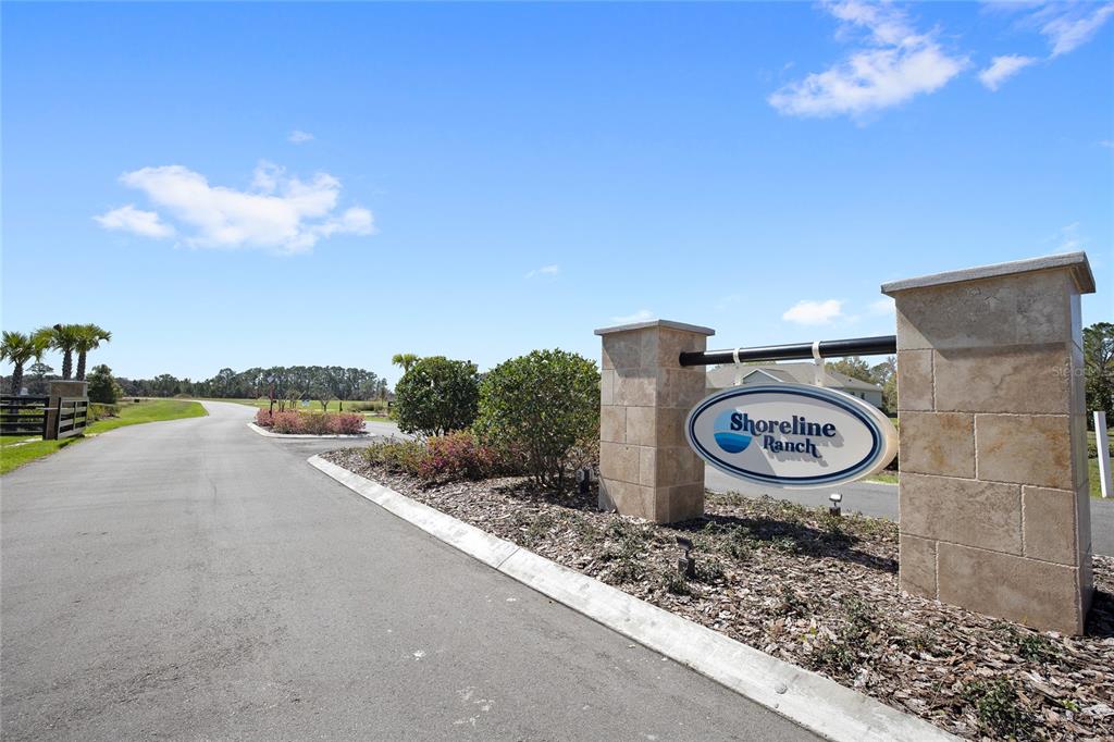 A stone entrance sign for Shoreline Ranch stands beside a paved road under a blue sky — 20280 SHORELINE RANCH LANE, UMATILLA, FL