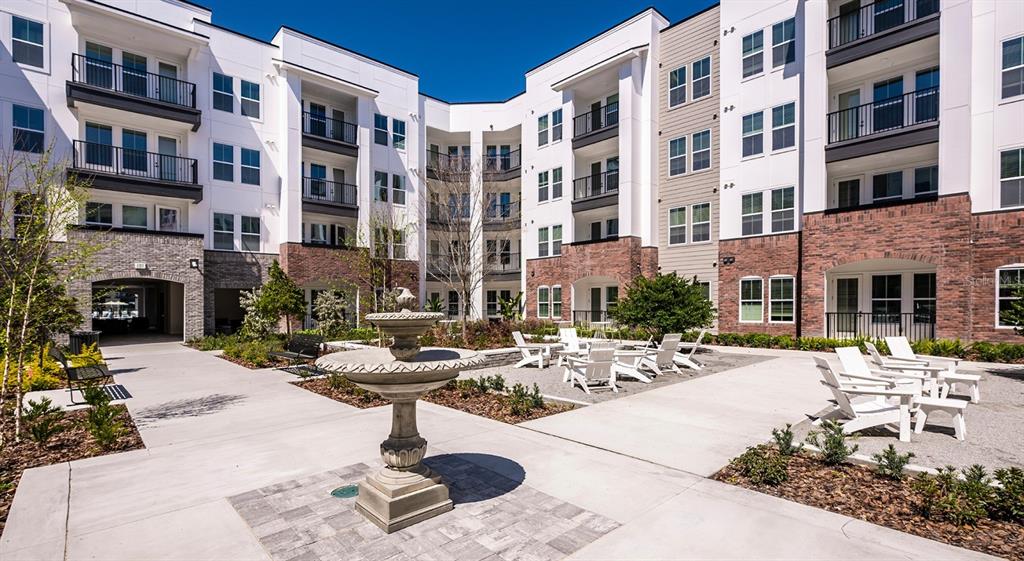 A courtyard with a stone fountain, patio seating, and modern apartment buildings under a blue sky — 700 AULTMAN STREET UNIT Broadway, KISSIMMEE, FL