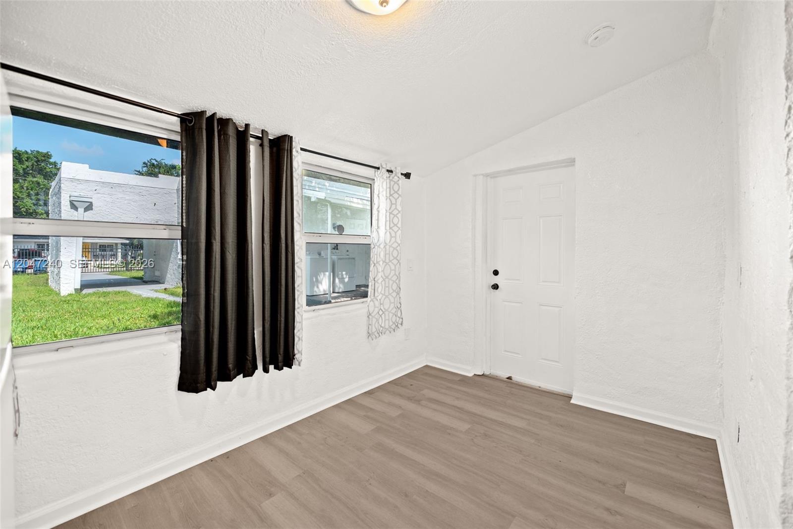 A room with white walls, wood-look flooring, a white door, and windows overlooking a yard — MIAMI, FL