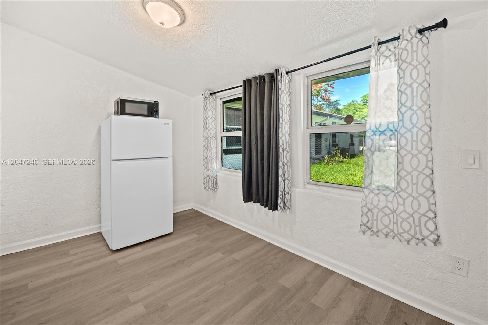 A room with light wood-look flooring, white walls, a refrigerator, and two windows with curtains — MIAMI, FL