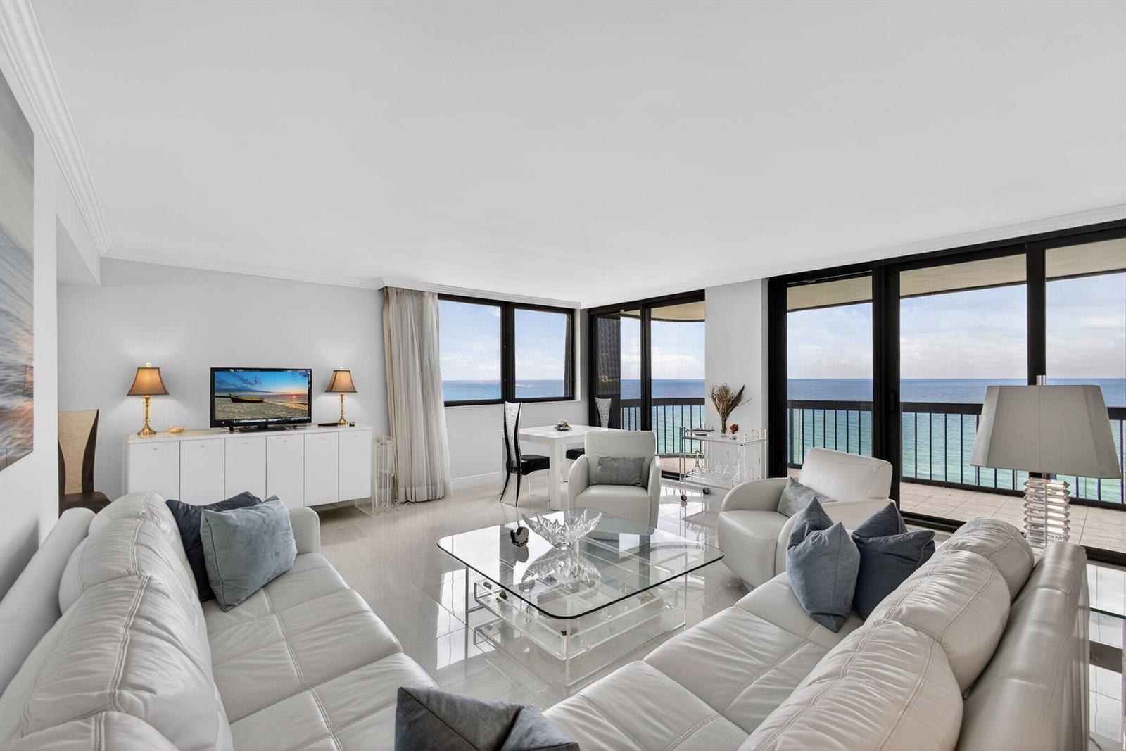 A bright living room features white leather sofas, a glass coffee table, and ocean views — 5380 N Ocean Drive # 12e, Singer Island FL 33404, SINGER ISLAND, FL