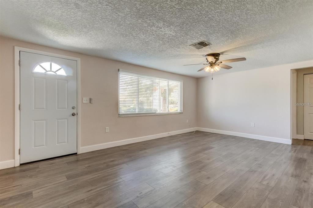 An empty living room features light-colored walls, wood-look flooring, a ceiling fan, and a front door — 7144 FAIRFAX DRIVE, PORT RICHEY, FL