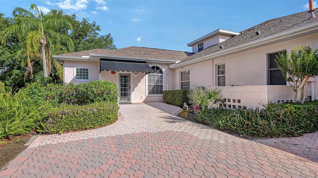 A paved driveway leads to the entrance of a light-colored house with a black awning — 7025 N SERENOA DRIVE, SARASOTA, FL