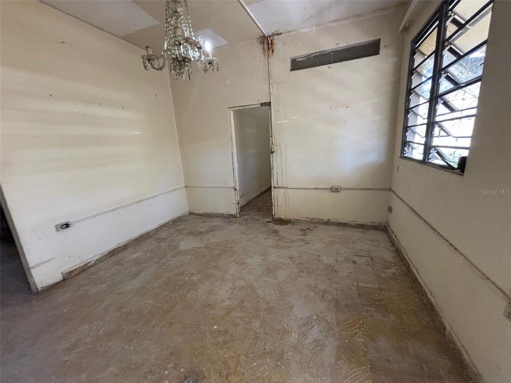 An empty room features a crystal chandelier, tiled flooring, and a window with security bars — D-13C Fajardo St. MIRAPALMERAS, SAN JUAN, PR