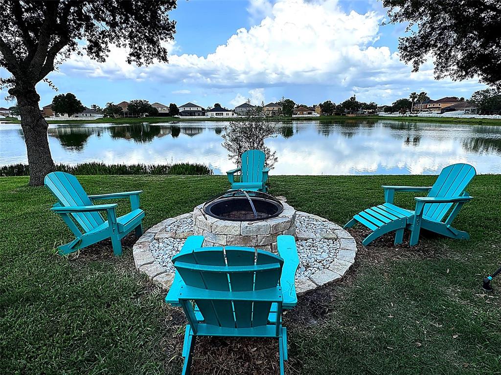 A stone fire pit with four turquoise Adirondack chairs sits on a lawn by a lake — 30913 BRIDGEGATE DRIVE, WESLEY CHAPEL, FL