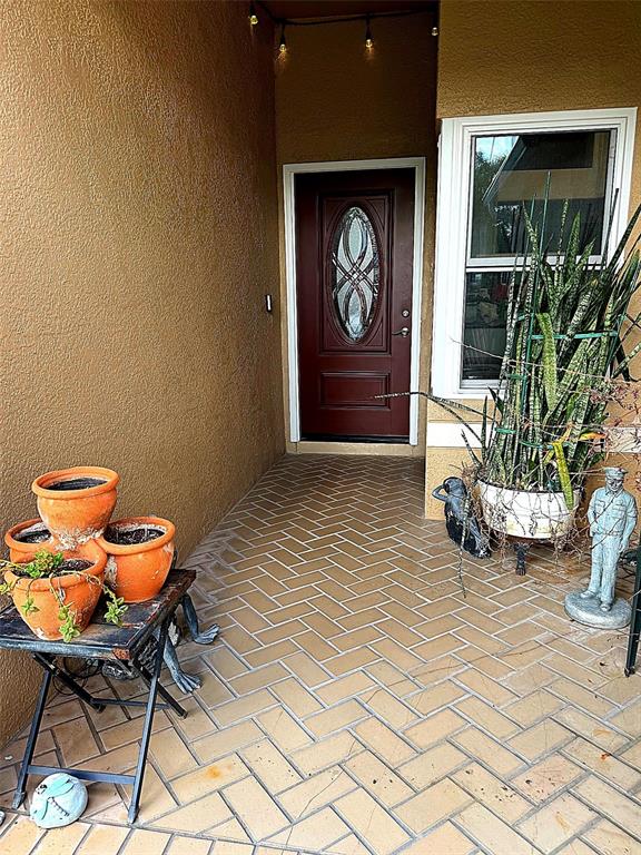 A covered porch features herringbone-patterned tile flooring, a dark red front door, and potted plants — 30913 BRIDGEGATE DRIVE, WESLEY CHAPEL, FL
