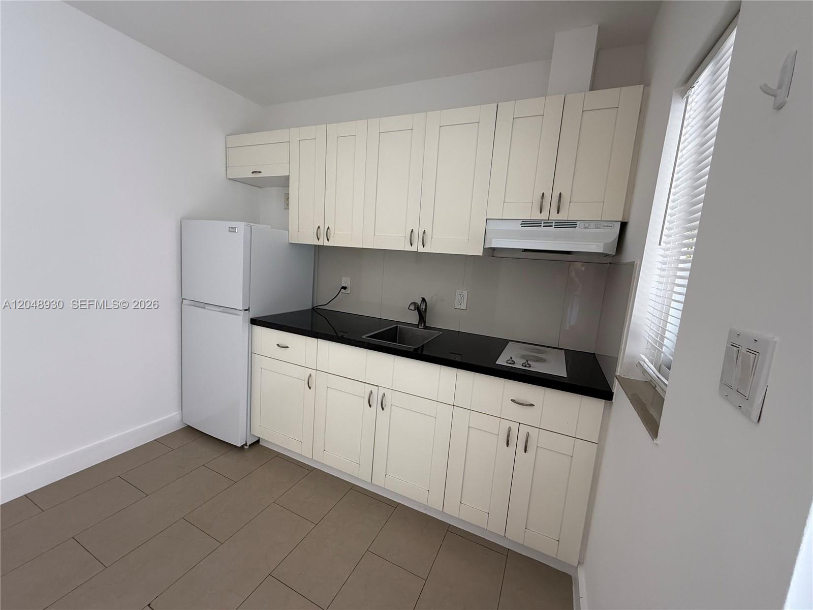 A small kitchen features white cabinets, black countertops, a white refrigerator, and tiled flooring — 242 NE 55th Ter # 2, Miami FL 33137, MIAMI, FL