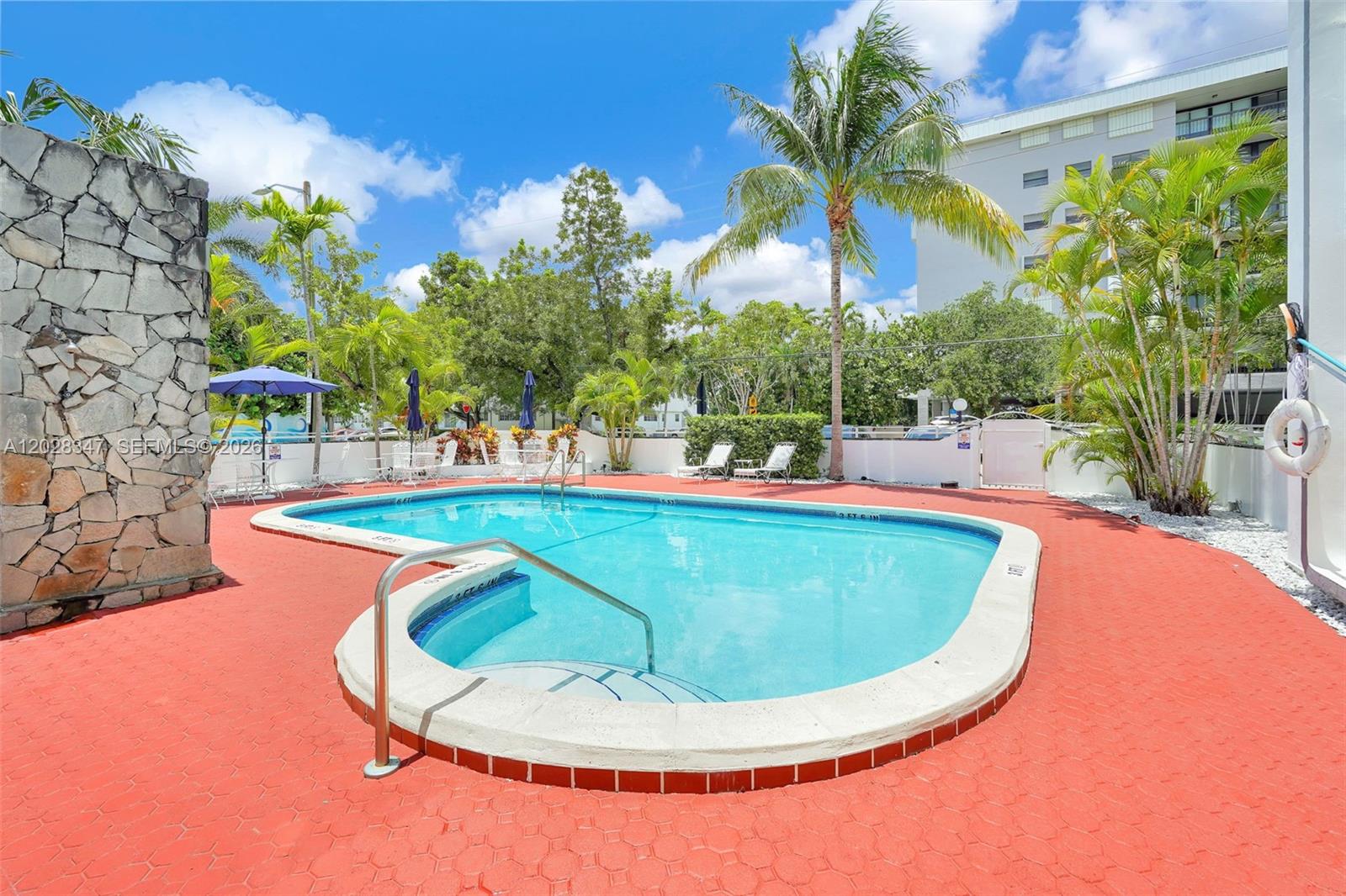 An outdoor swimming pool with a red deck is surrounded by palm trees and landscaping — 1075 93rd St # 207, Bay Harbor Islands FL 33154, BAY HARBOR ISLANDS, FL