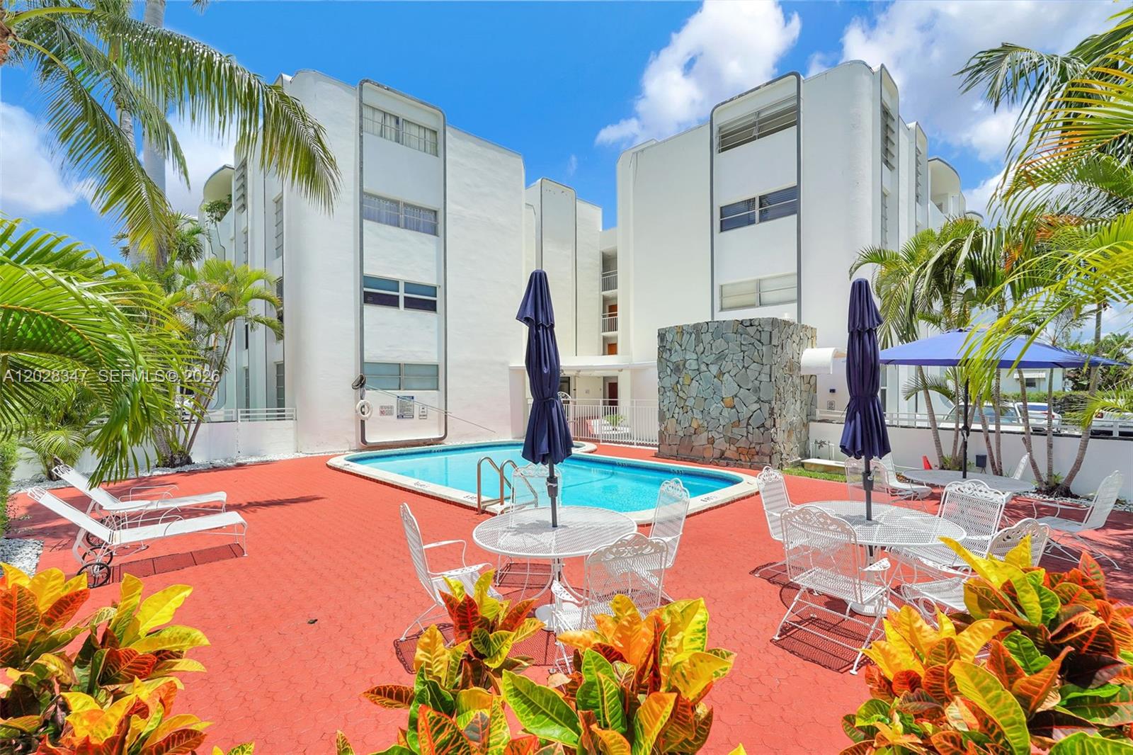 An outdoor swimming pool area with lounge chairs, tables, and umbrellas in front of apartments — 1075 93rd St # 207, Bay Harbor Islands FL 33154, BAY HARBOR ISLANDS, FL