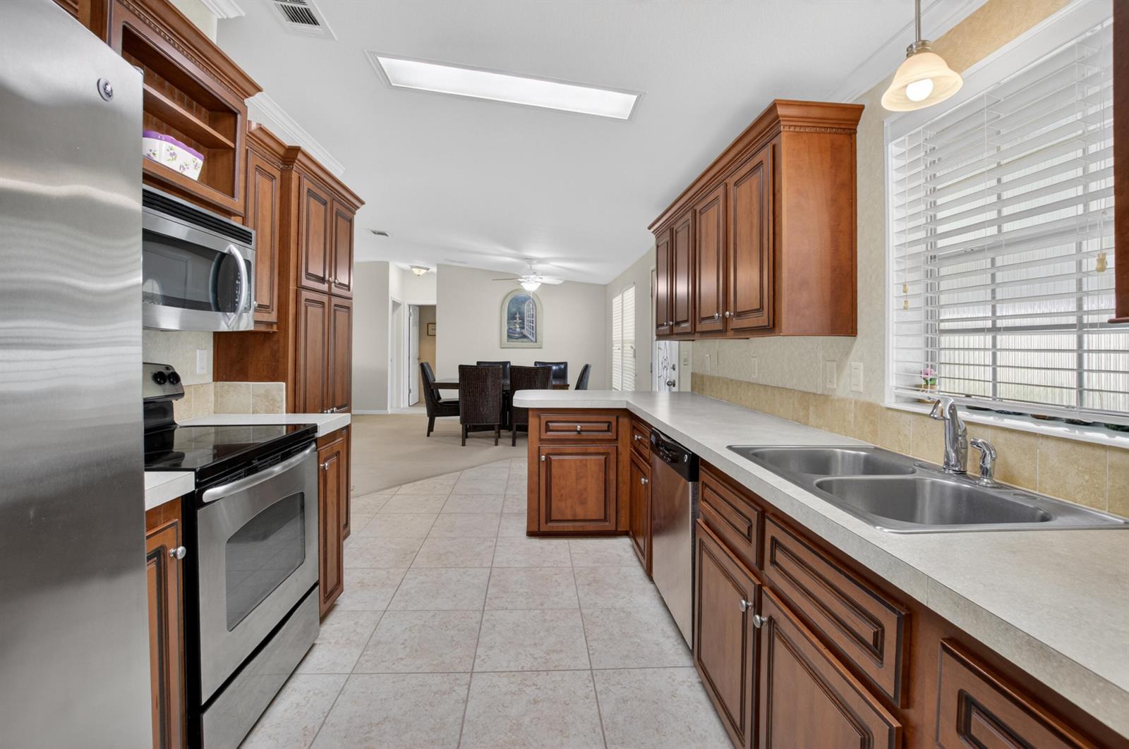 A kitchen with wood cabinets, stainless steel appliances, and tile flooring leads to a dining area — 5006 Curacas Bay, Boynton Beach FL 33436, BOYNTON BEACH, FL
