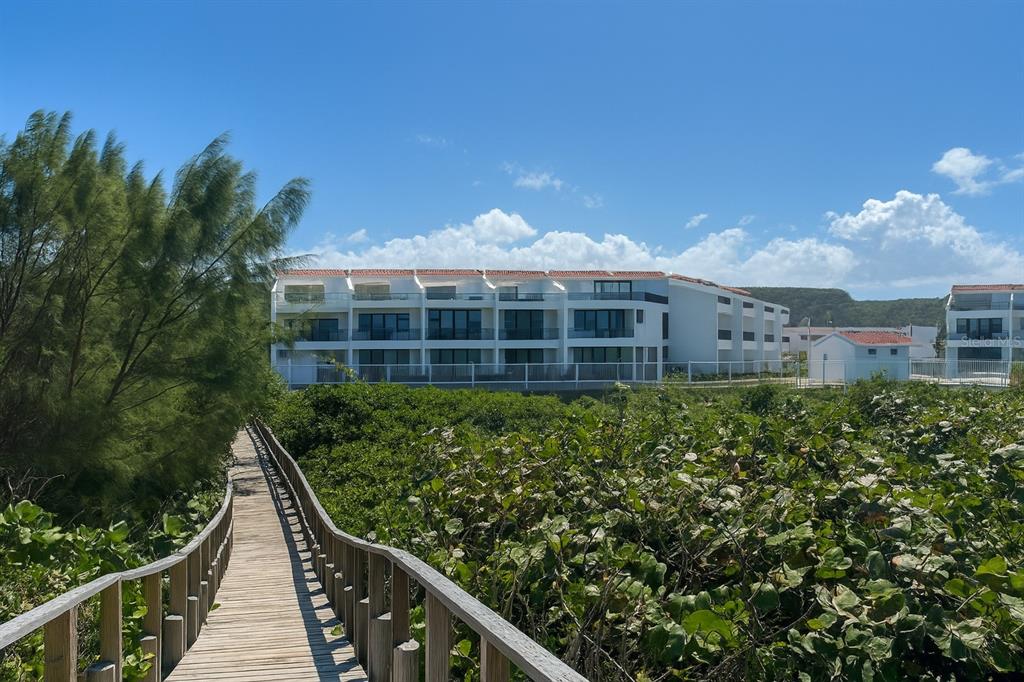 A wooden boardwalk leads through coastal vegetation toward a multi-story white apartment building — C-116 MARBELA CASA DE PLAYA UNIT 1, ISABELA, PR