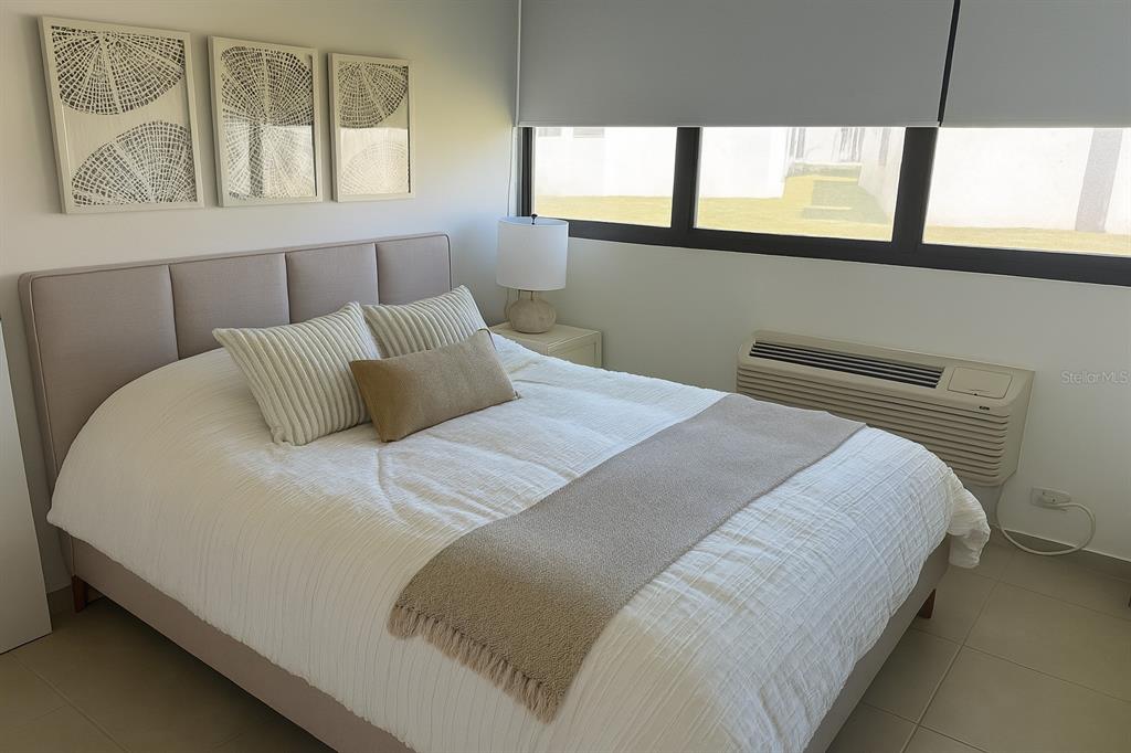 A bedroom features a beige upholstered bed, white bedding, wall art, and a window unit — C-116 MARBELA CASA DE PLAYA UNIT 1, ISABELA, PR