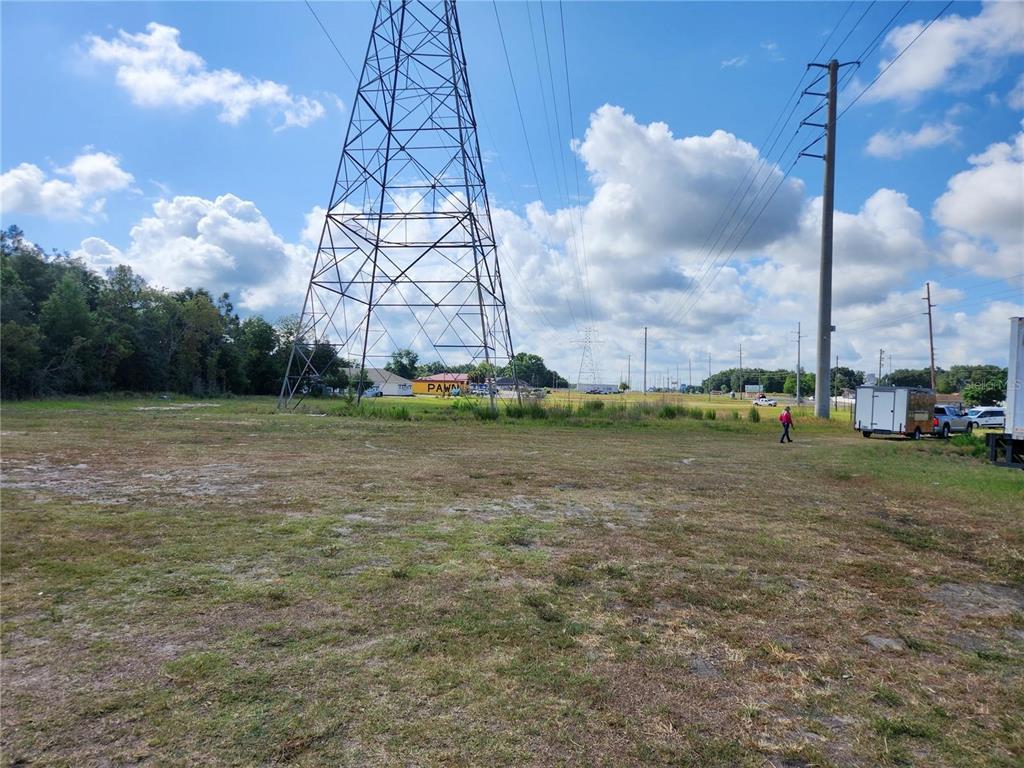 A large, grassy vacant lot features a tall metal electrical transmission tower under a cloudy sky — 7013 SE MARICAMP ROAD, OCALA, FL