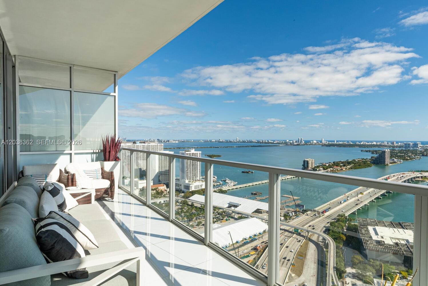 A high-rise balcony features outdoor seating overlooking a bay, a bridge, and city buildings — 1100 Biscayne Blvd # 4103, Miami FL 33132, MIAMI, FL