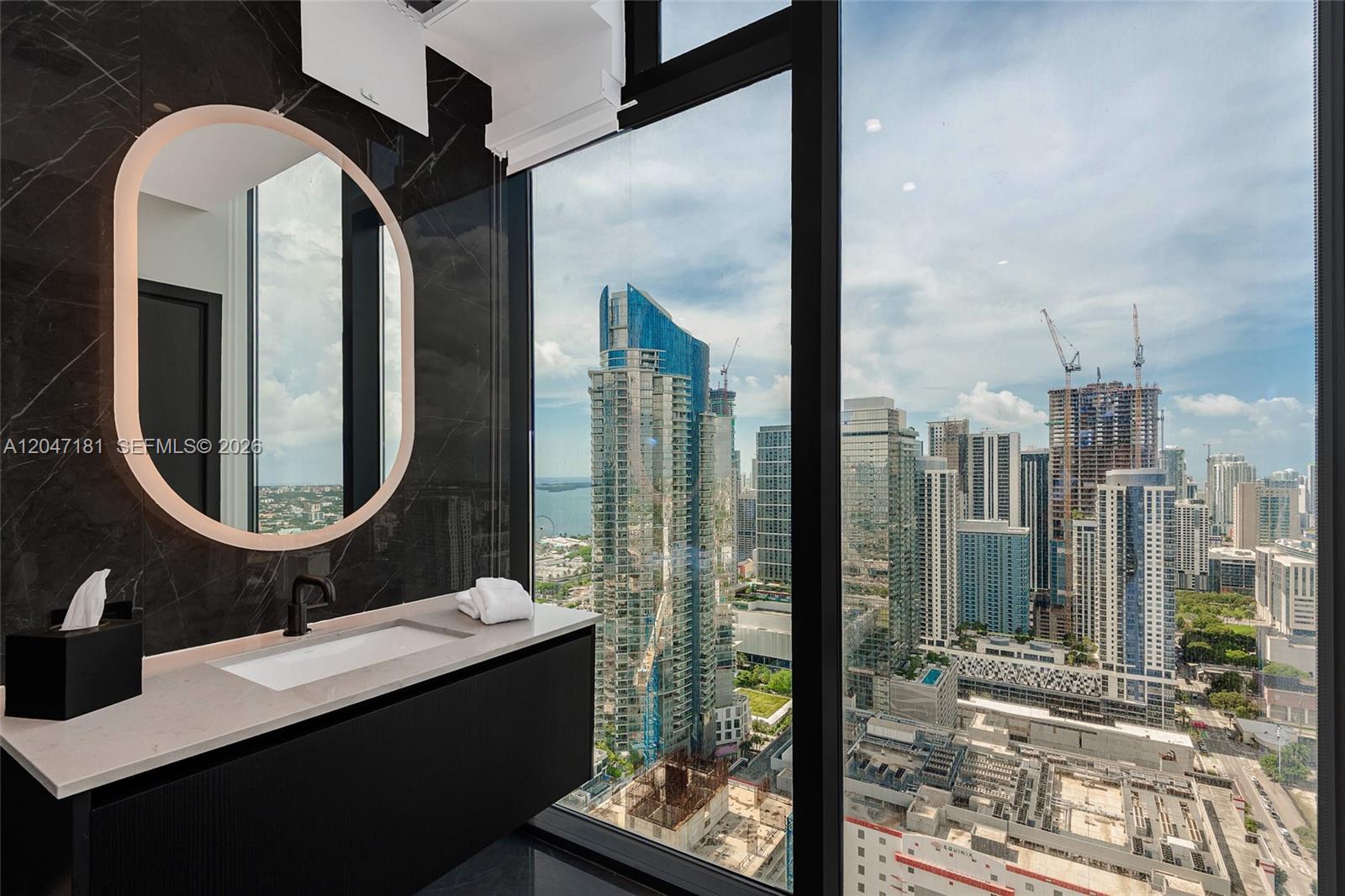 A modern bathroom features a floating vanity, round backlit mirror, and floor-to-ceiling city skyline views — 20 NE 11 # 4404, Miami FL 33132, MIAMI, FL