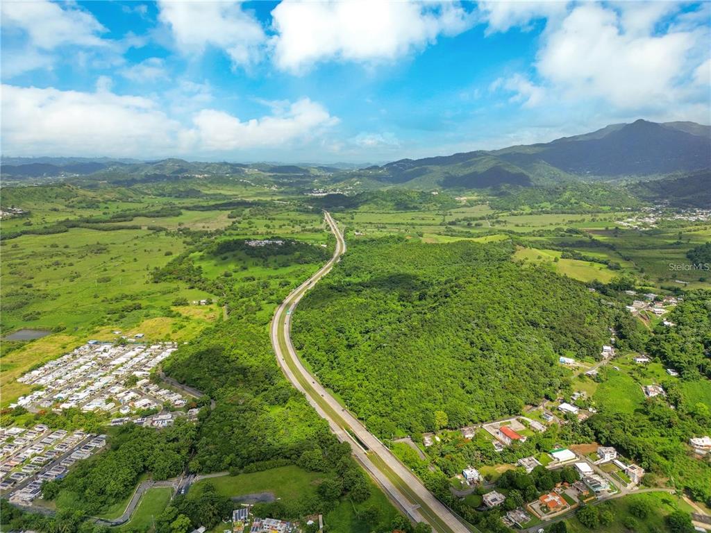 An aerial view shows a highway winding through a lush, mountainous landscape with residential areas — BO MAIZALES CALLE BORGES FINAL, NAGUABO, PR