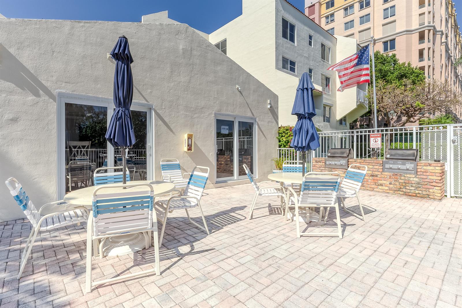 A paved outdoor patio features two dining tables with blue umbrellas and chairs near grills — 2850 NE 14th St Causeway # 308b, Pompano Beach FL 33062, POMPANO BEACH, FL