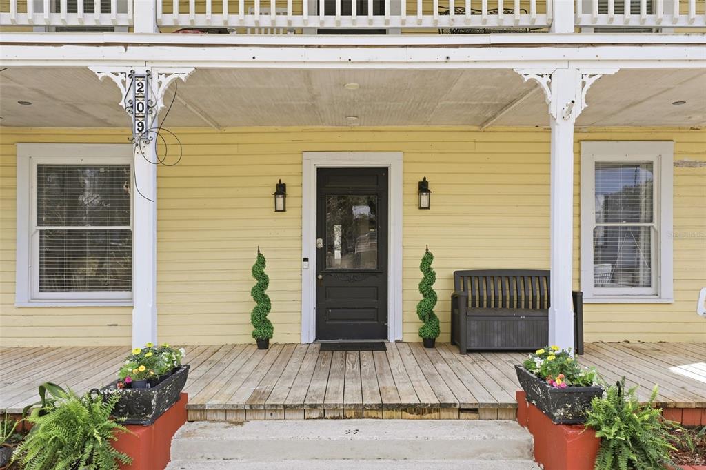 The front porch of a yellow house features a black door, wooden decking, and planters — 209 E 5TH STREET, SANFORD, FL