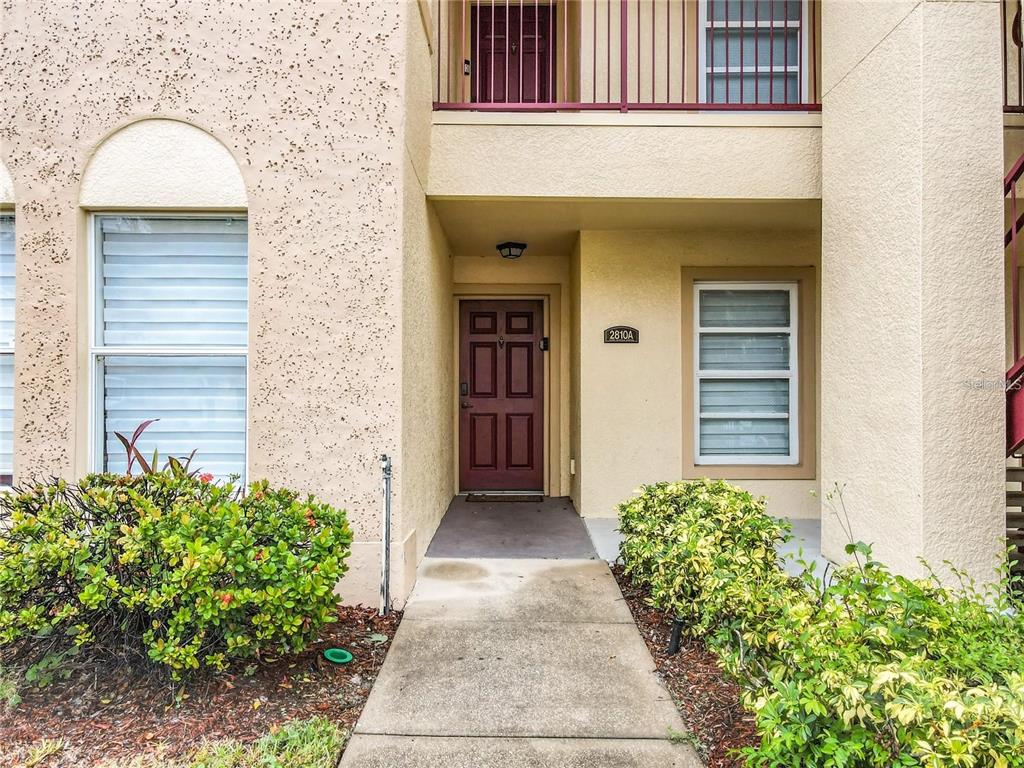 A concrete walkway leads to the maroon front door of a tan stucco apartment building — 2810 CLUB CORTILE CIRCLE UNIT A, KISSIMMEE, FL