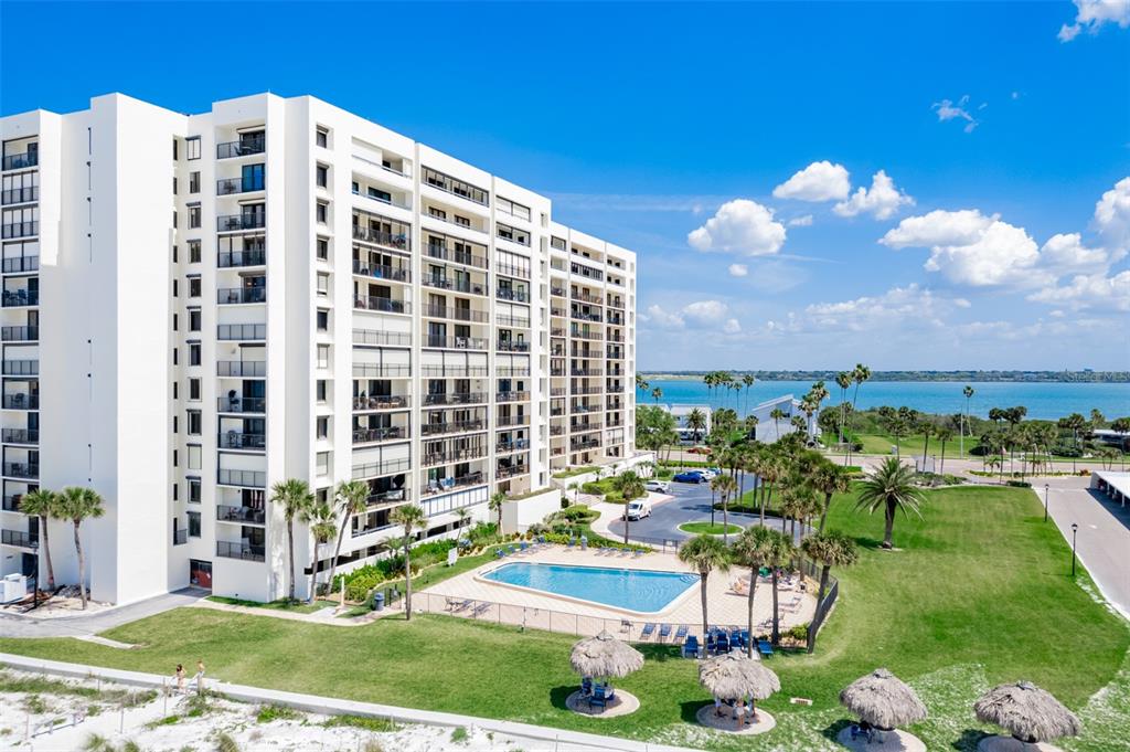 An aerial view shows a multi-story white apartment building with a pool and beach access — 1460 GULF BOULEVARD UNIT 1205, CLEARWATER BEACH, FL