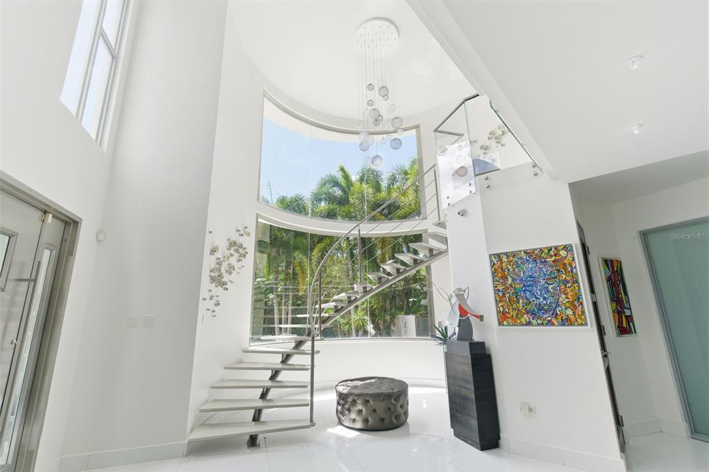 A modern foyer features a curved glass wall, a spiral staircase, and white tile flooring — 57 QUINTAS DE SANTA MARIA II, MAYAGUEZ, PR