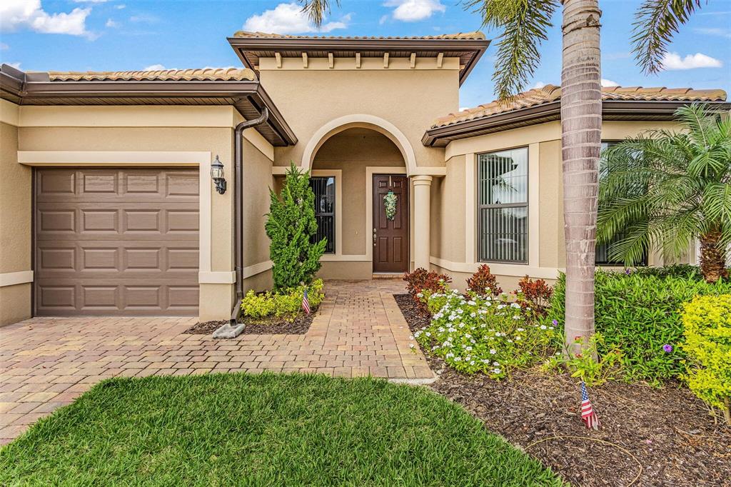 A single-story house exterior features a tan stucco facade, a brown garage door, and landscaping — 12437 GOLDEN SAGE DRIVE, SARASOTA, FL