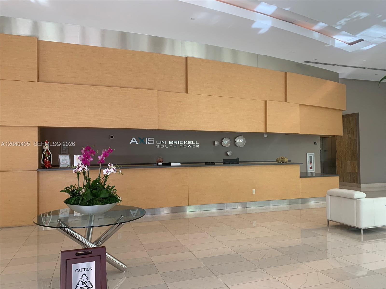 A modern lobby features a long wooden reception desk, a glass table with orchids, and tile flooring — 79 SW 12th St, Miami FL 33130, MIAMI, FL