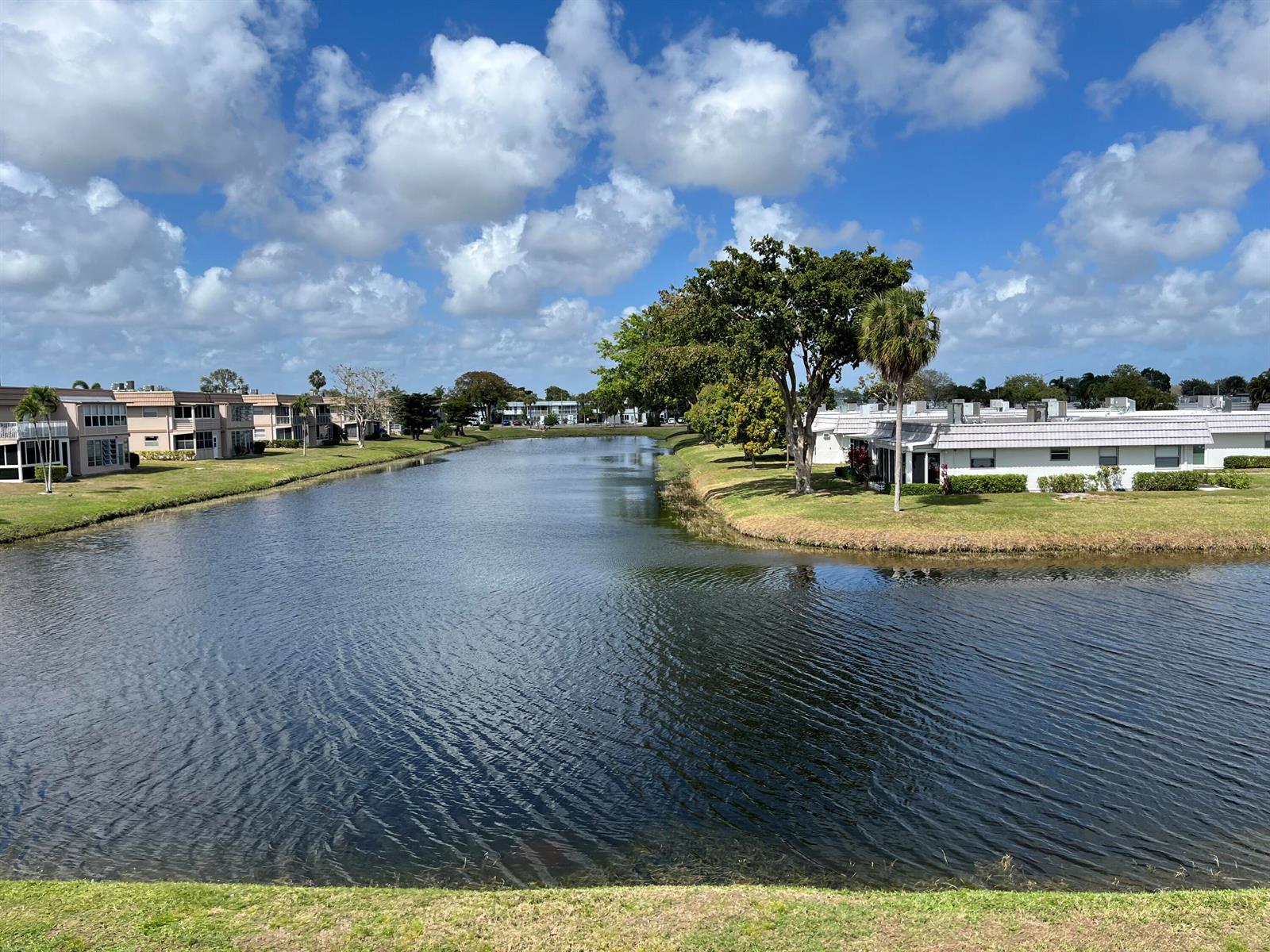 A canal flows between residential buildings and green lawns under a blue sky with clouds — 192 Monaco D, Delray Beach FL 33446, DELRAY BEACH, FL