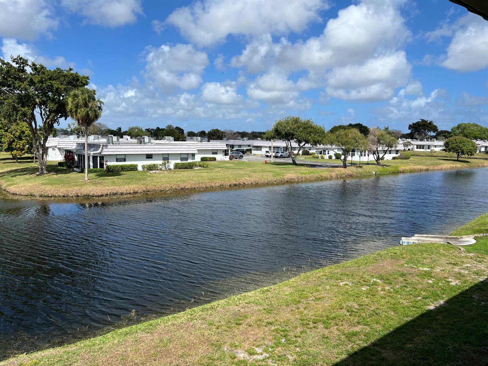 A view of a canal with white residential buildings and trees under a blue sky — 192 Monaco D, Delray Beach FL 33446, DELRAY BEACH, FL