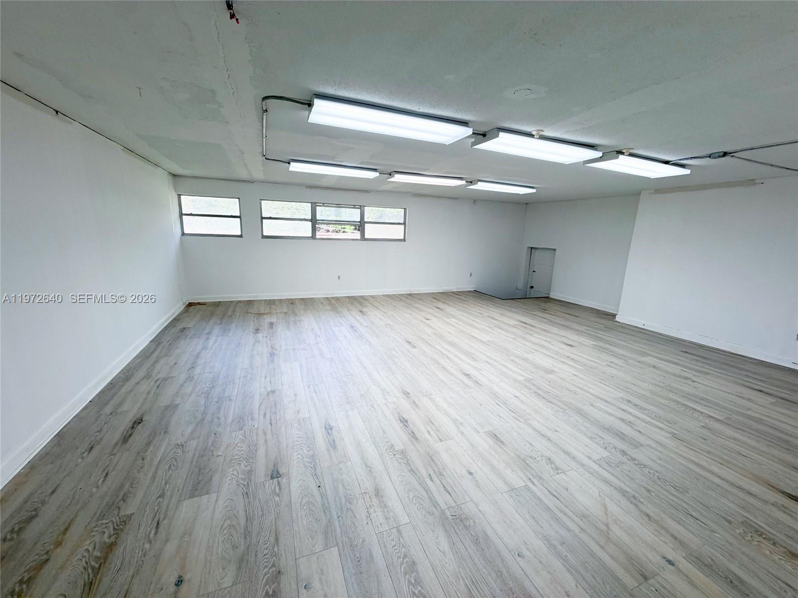 An empty room features light wood-look flooring, white walls, and surface-mounted fluorescent lighting fixtures — 18500 NE 5th Ave, Miami FL 33179, MIAMI, FL