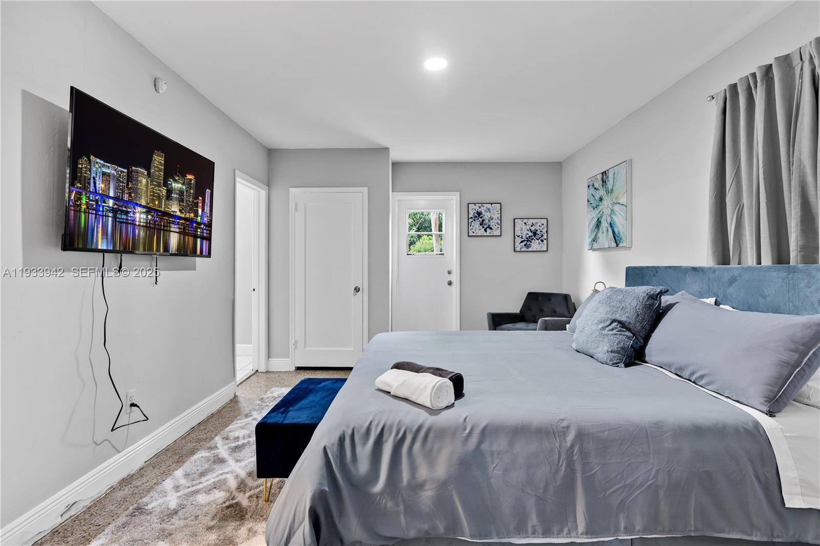 A bedroom features a bed with blue bedding, a wall-mounted television, and gray carpeting — 427 NE 107th St # 2, Miami FL 33161, MIAMI, FL