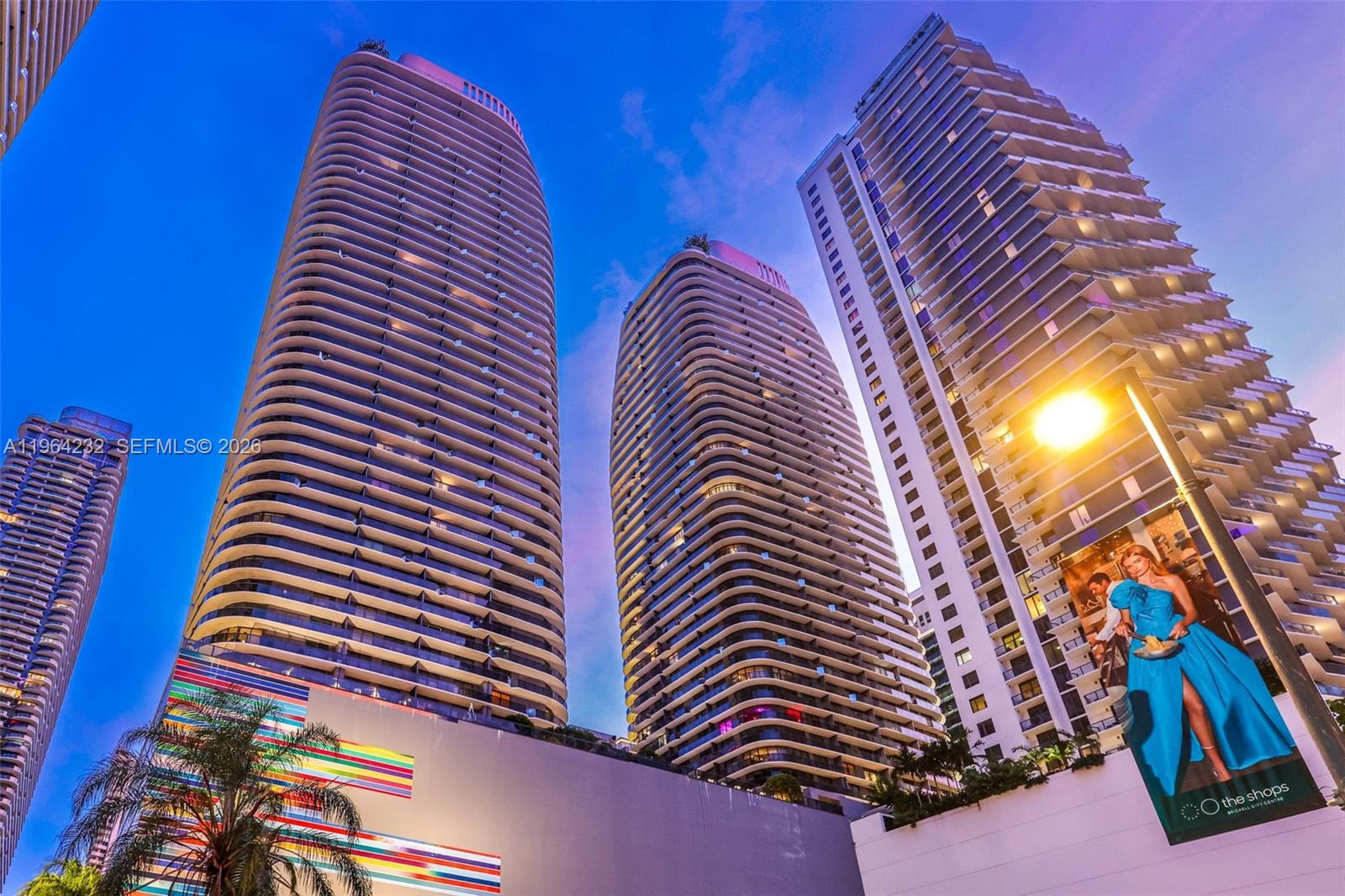 A low-angle view of three modern high-rise residential towers against a blue twilight sky — 55 SW 9th St # 3906, Miami FL 33130, MIAMI, FL