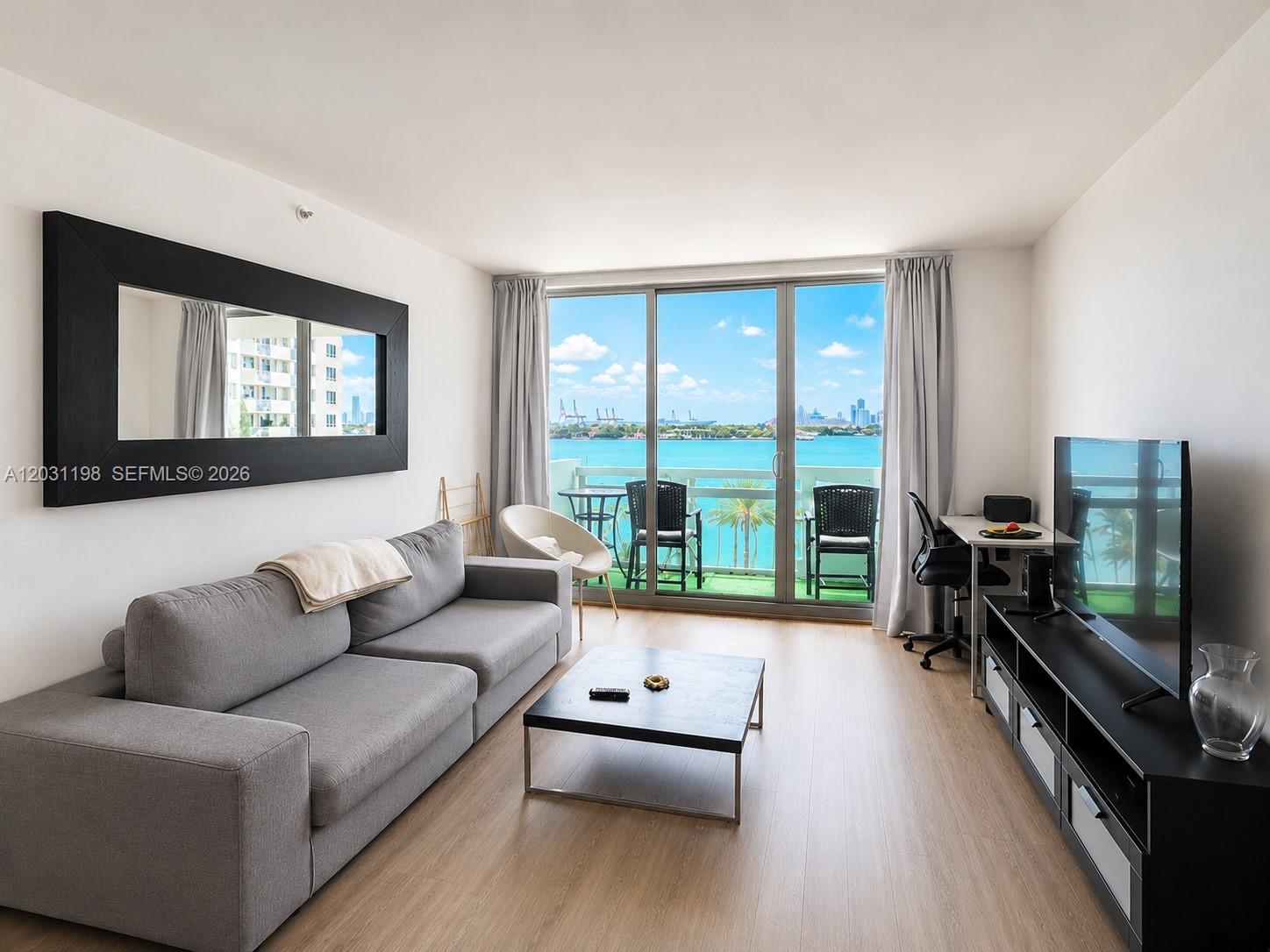 A living room features a gray sofa, black coffee table, TV console, and a balcony view — 1500 Bay Rd # 718S, Miami Beach FL 33139, MIAMI BEACH, FL