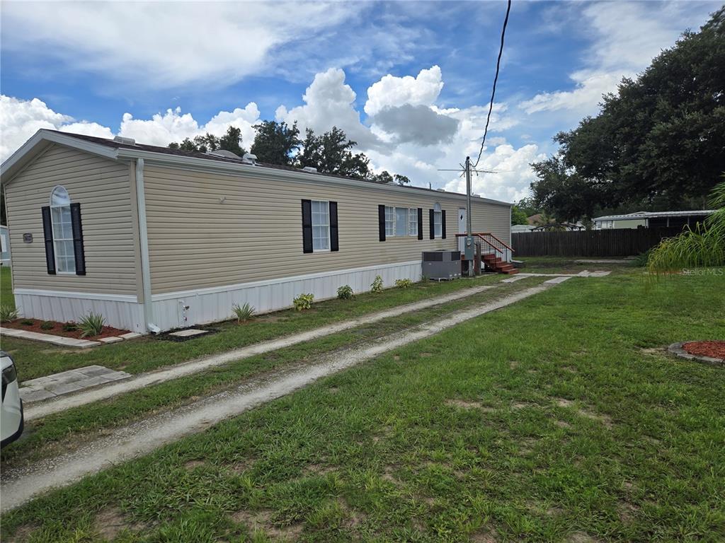 A tan mobile home with white skirting sits on a grassy lot with a gravel driveway — 16127 NARROW STREET, BROOKSVILLE, FL
