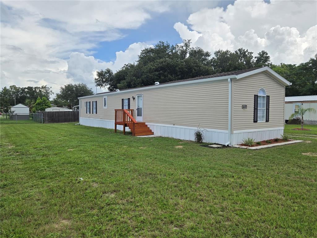 A beige manufactured home with a wooden deck stands on a grassy lot under clouds — 16127 NARROW STREET, BROOKSVILLE, FL