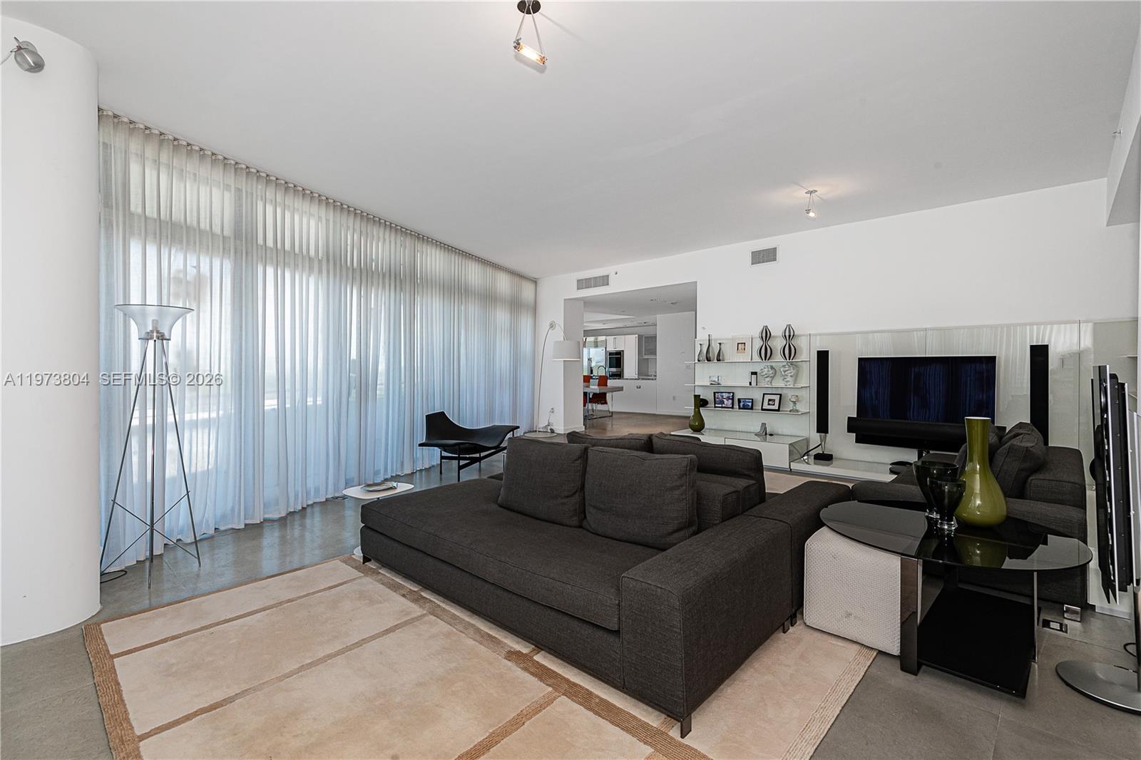 A modern living room features a dark gray sofa, a beige area rug, and floor-to-ceiling curtains — 6101 Aqua Ave # 702, Miami Beach FL 33141, MIAMI BEACH, FL