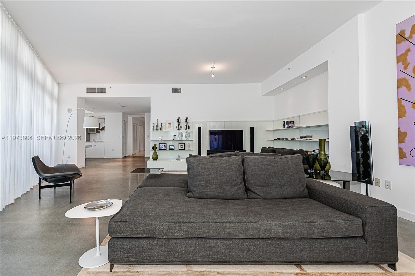 A modern living room features a large gray sofa, concrete floors, and white walls — 6101 Aqua Ave # 702, Miami Beach FL 33141, MIAMI BEACH, FL
