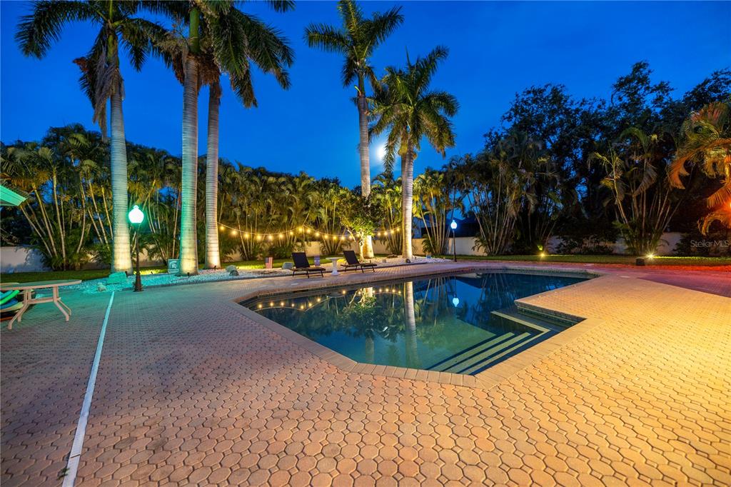A swimming pool with a brick paver deck is surrounded by palm trees at night — 4930 SANDPIPER LANE S, ST PETERSBURG, FL