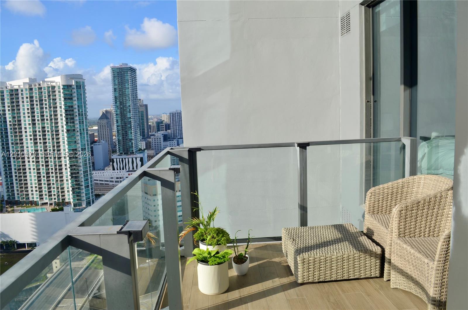 A balcony with a wicker chair, ottoman, potted plants, and a city skyline view — 68 SE 6th St # 2708, Miami FL 33131, MIAMI, FL