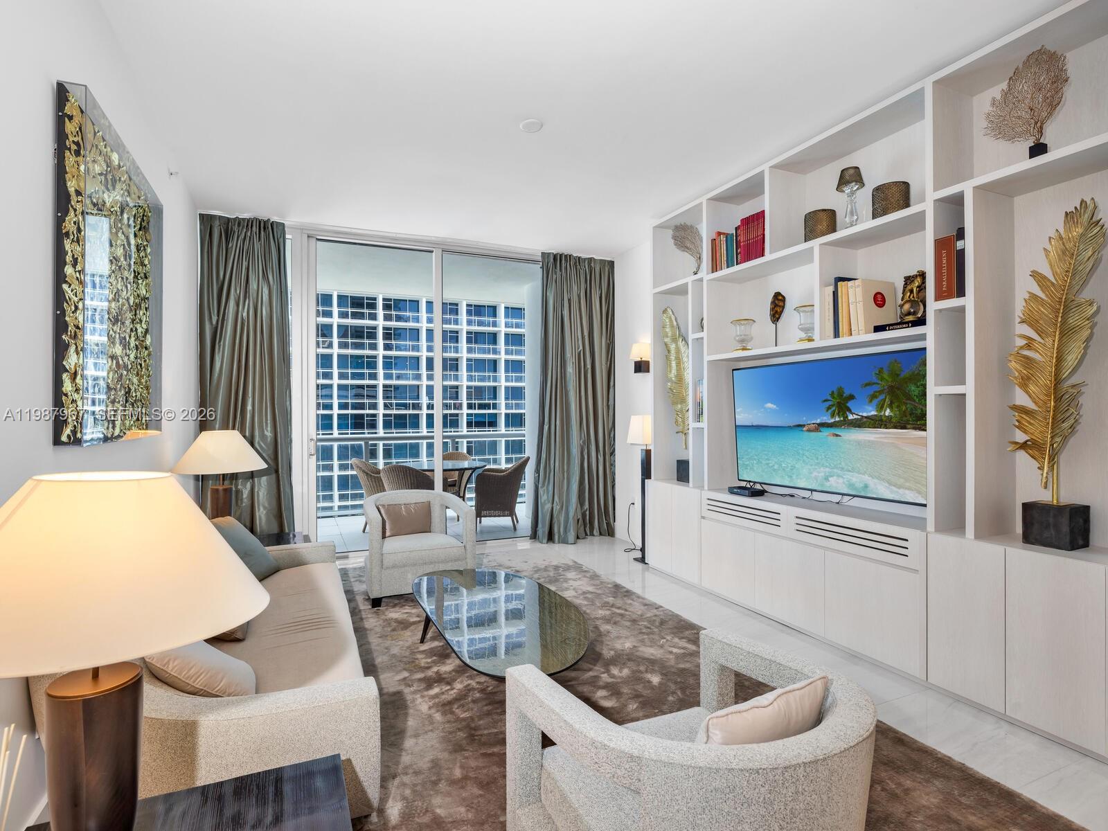 A modern living room features a large built-in shelving unit, a television, and balcony access — 6799 Collins Ave # 501, Miami Beach FL 33141, MIAMI BEACH, FL