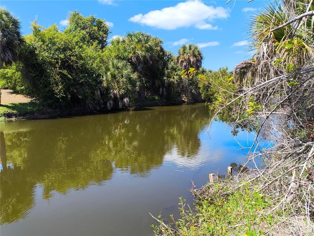 A narrow canal flows past a lush, tree-lined bank under a bright blue sky — 12289 CLARENDON AVENUE, PORT CHARLOTTE, FL