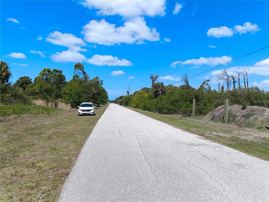 A paved road stretches into the distance between grassy areas and dense green vegetation — 12289 CLARENDON AVENUE, PORT CHARLOTTE, FL