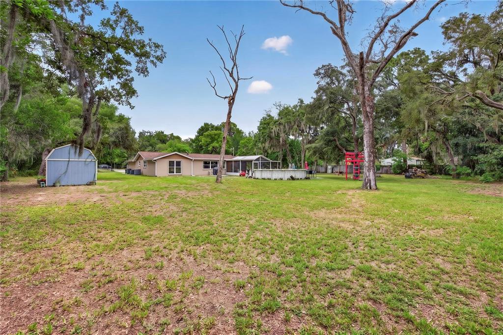 A backyard view of a single-story house with a shed, pool, and large trees — 13129 FILBERT STREET, HUDSON, FL